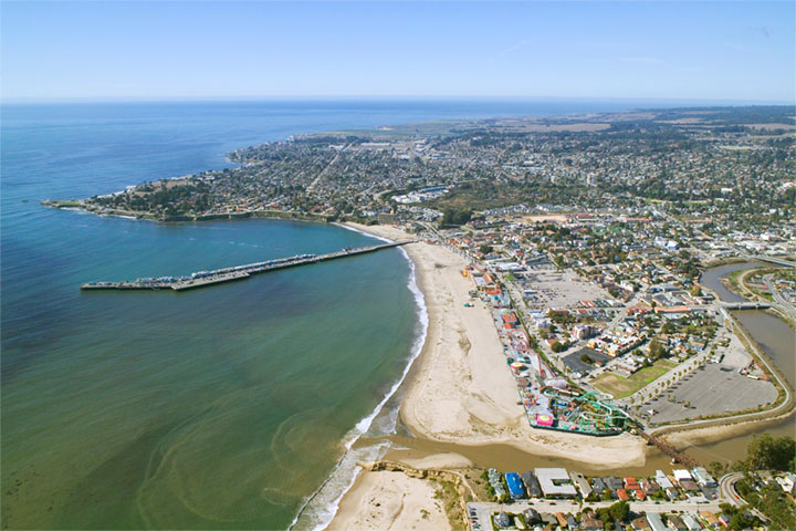 Aerial view of a coastal town with pier, beach, and boardwalk area next to blue ocean, sunny day, blue and tan dominant